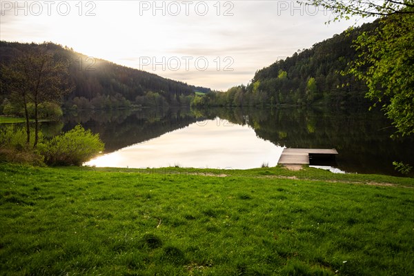 A lake in a landscape shot. A sunset and the natural surroundings are reflected in the water of the reservoir. Marbach reservoir, Odenwald, Hesse