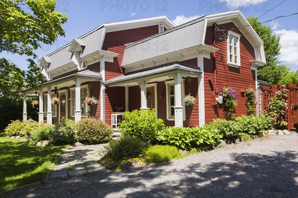 Old 1892 burgundy with white trim pinewood plank house facade with mansarde style grey sheet metal roof and landscaped front yard in summer, Quebec, Canada, North America