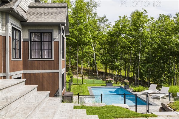 In-ground swimming pool and rear view of contemporary natural stone and brown stained wood and cedar shingles clad luxurious bungalow in summer, Quebec, Canada, North America