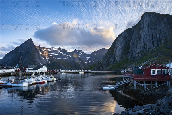 The village of Hamnoy. On the right, a typical red wooden house (rorbuer) on wooden stilts. On the left, other houses and boats in a small harbour. In the background the mountain Olstinden. At night at the time of the midnight sun in good weather, some clouds in the sky. Early summer. Hamnoy, Moskenesoya, Lofoten, Norway, Europe