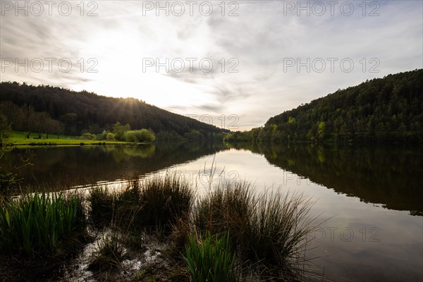 A lake in a landscape shot. A sunset and the natural surroundings are reflected in the water of the reservoir. Marbach reservoir, Odenwald, Hesse