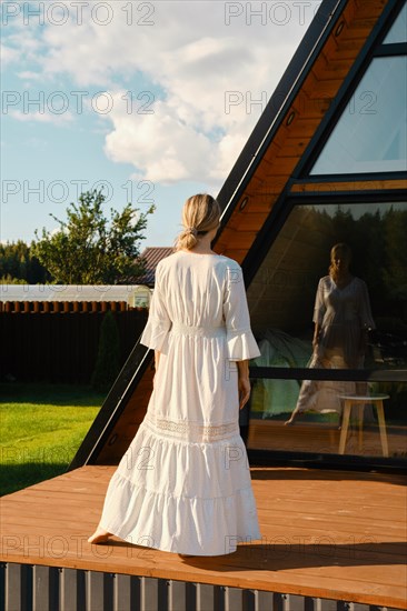 Barefoot woman standing on wooden terrace of tiny house and looking at her reflection in large window