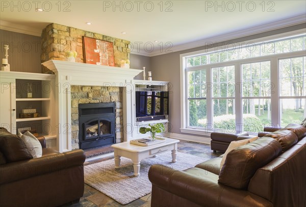 Brown leather sofa, sitting chairs and artificial stone fireplace in the living room with earth tone slate flooring inside a contemporary cottage style home, Quebec, Canada, North America