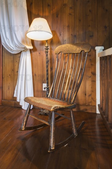 Spindle high back antique wooden rocking chair and lit pedestal lamp in hallway on upper floor inside a New Hampton style home, Quebec, Canada, North America
