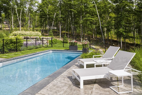 Two white long chairs on edge of in-ground swimming pool enclosed by clear glass and black metal fence in residential backyard in summer, Quebec, Canada, North America