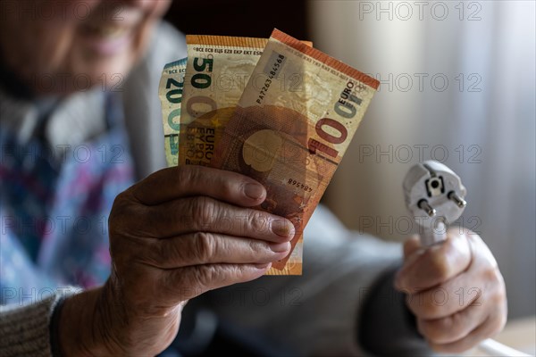 Senior citizen holding money and a power cable with plug in her hand at home, symbolising energy costs and poverty, Cologne, North Rhine-Westphalia, Germany, Europe