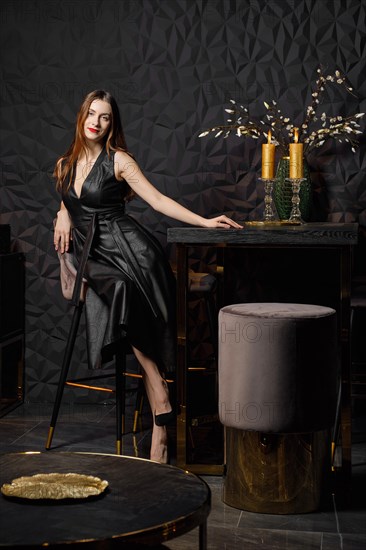Happy girl in faux leather dress sitting in chair near bar table at home