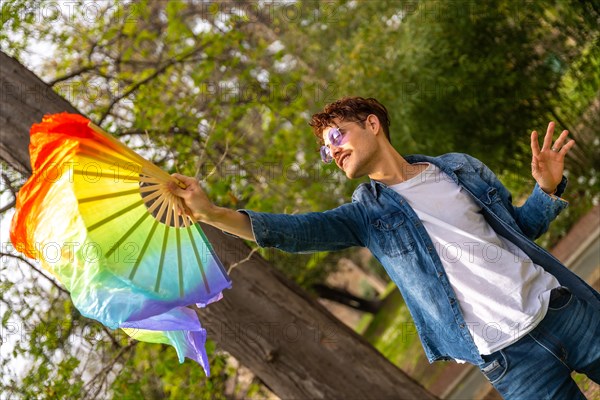 Dynamic shot of a happy casual gay male artist moving a rainbow fan dancing in a park