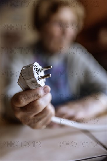 Senior citizen holding a power cable with plug in her hand at home, symbolising energy costs and poverty, Cologne, North Rhine-Westphalia, Germany, Europe