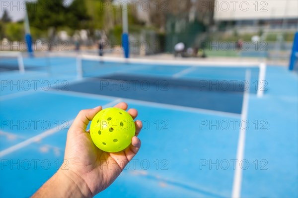 Unrecognizable person holding a plastic yellow pickleball ball with blue outdoor court on the background