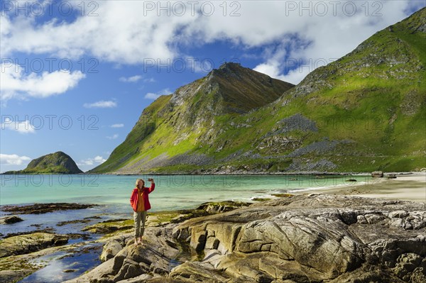Landscape with sea at the sandy beach of Haukland (Hauklandstranda) with the mountain Veggen. A woman stands barefoot on a rock and takes a selfie with her mobile phone. Good weather, blue sky and a few clouds. Early summer. Haukland Beach, Haukland, Vestvagoya, Lofoten, Norway, Europe