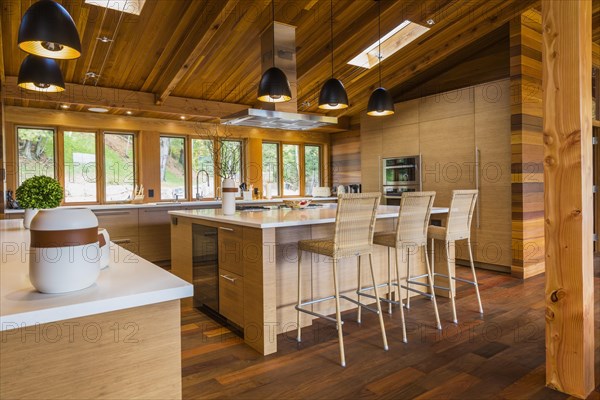 Bamboo wood buffet and island with white quartz countertops and high wicker chairs, cabinets in kitchen with Ipe wood floor plus black industrial style pendant lighting fixtures inside luxurious stained cedar and timber wood home with panoramic windows, Quebec, Canada, North America
