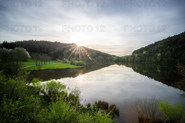 A lake in a landscape shot. A sunset and the natural surroundings are reflected in the water of the reservoir. Marbach reservoir, Odenwald, Hesse