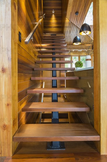 Looking up illuminated wooden staircase with opened steps and clear glass railing leading to upstairs floor inside luxurious stained cedar and timber wood home, Quebec, Canada, North America