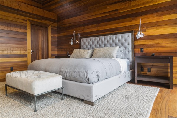 King size bed with grey upholstered panel headboard, ottoman and wooden end tables in master bedroom with grey nuanced rug and Ipe wood floor inside luxurious stained cedar and timber wood home, Quebec, Canada, North America