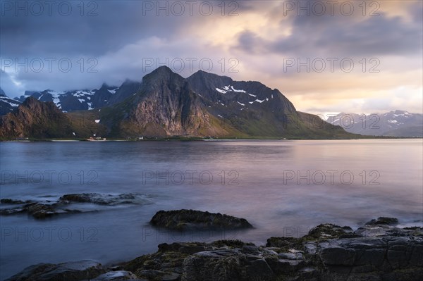 Landscape with sea and mountains on the Lofoten Islands, view across the fjord to the small town of Flakstad and the mountain Flakstadtinden as well as other mountains. Rocks in the foreground. At night at the time of the midnight sun in good weather. Some clouds in the sky, some sunlight on the mountains. Long exposure, blurred clouds. Early summer. Flakstadoya, Lofoten, Norway, Europe