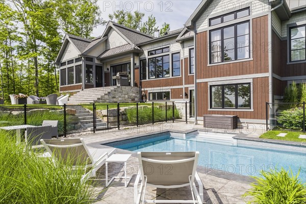 In-ground swimming pool and rear view of contemporary natural stone and brown stained wood and cedar shingles clad luxurious bungalow style home in summer, Quebec, Canada, North America