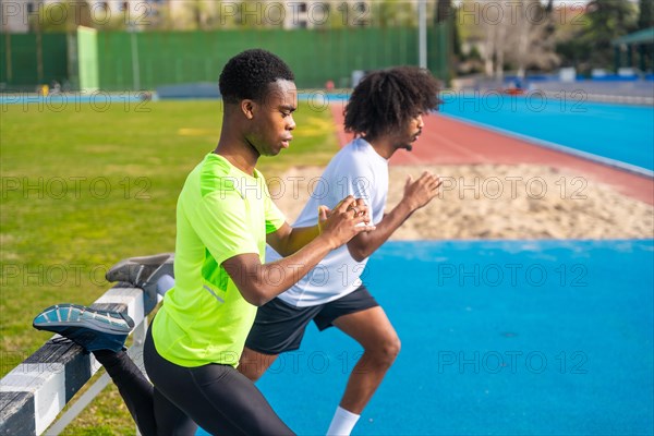 Two young african american runners warming up stretching legs in an outdoor track using a fence