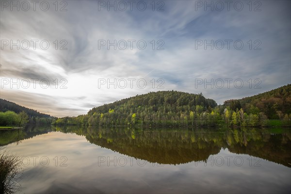 A lake in a landscape shot. A sunset and the natural surroundings are reflected in the water of the reservoir. Marbach reservoir, Odenwald, Hesse