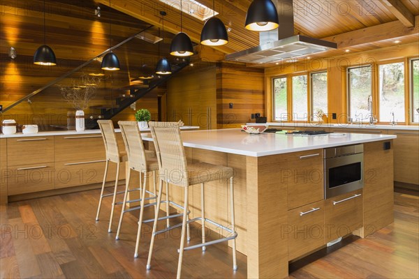 Bamboo wood island with white quartz countertops and high wicker chairs, buffet in kitchen with Ipe wood floor plus black industrial style pendant lighting fixtures inside luxurious stained cedar wood and timber home with panoramic windows, Quebec, Canada, North America