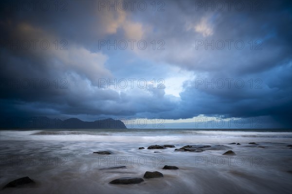 On the beach at Vikten. The sea washes around some rocks. Mountains in the background. At night at the time of the midnight sun. Cloudy, dramatic sky. Early summer. Long exposure. Vikten, Flakstadoya, Lofoten, Norway, Europe
