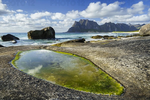 Seascape on the beach at Uttakleiv (Utakleiv), in the foreground rocks and a rocky outcrop filled with water. In the background the mountain Hogskolmen. Sun and clouds. Early summer. Uttakleiv, Vestvagoya, Lofoten, Norway, Europe
