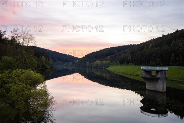 A lake in a landscape shot. A sunset and the natural surroundings are reflected in the water of the reservoir. Marbach reservoir, Odenwald, Hesse