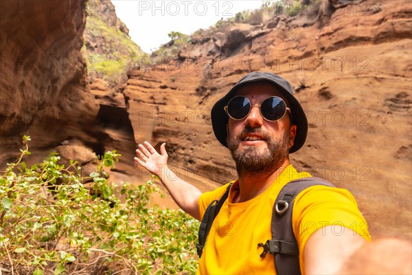 A man in a yellow shirt and sunglasses is taking a selfie in front of a rocky cliff. Concept of adventure and exploration, as the man is posing in a natural setting with a backpack on