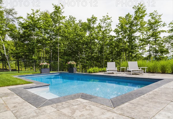 Two white long chairs on edge of in-ground swimming pool with flowers in planters in residential backyard in summer, Quebec, Canada, North America