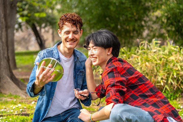 Gay couple making up using hand mirror sitting together in a park