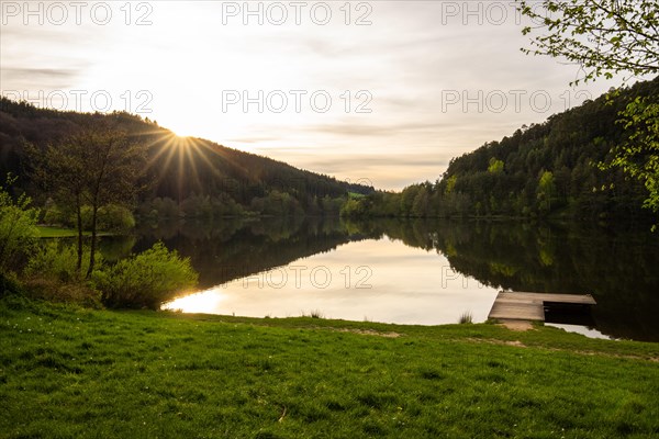 A lake in a landscape shot. A sunset and the natural surroundings are reflected in the water of the reservoir. Marbach reservoir, Odenwald, Hesse