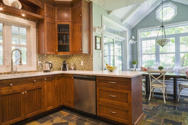 Kitchen with cherry wood cabinets, earth tone slate flooring and breakfast table with sitting bench and rustic chairs inside a contemporary cottage style home, Quebec, Canada, North America