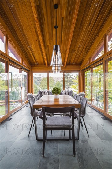 Four season gazebo and illuminated industrial style black with clear glass pendant lighting fixtures over wood and black metal dining table with grey upholstered chairs and heated slate floor inside luxurious stained cedar and timber wood home with panoramic windows, Quebec, Canada, North America