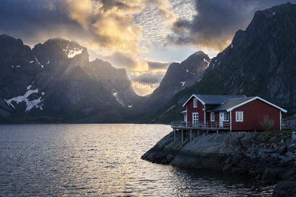 A typical red wooden house on wooden stilts (rorbuer) in the small village of Toppoya (Toppoy), overlooking the sea and the mountains. At night at the time of the midnight sun. Clouds and blue sky, the sun shines on a mountain peak. Toppoya, Moskenesoya, Lofoten, Norway, Europe