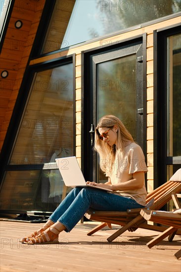 Woman checking mail on laptop sitting in a folding wooden chair on the terrace of summer house