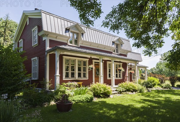 Old 1892 burgundy with white trim pinewood plank house facade with mansarde style grey sheet metal roof and landscaped front yard in summer, Quebec, Canada, North America