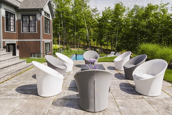 White and grey cloth covered sitting chairs on backyard patio and rear view of contemporary natural stone and brown stained wood and cedar shingles clad luxurious bungalow style home in summer, Quebec, Canada, North America