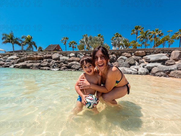 A woman and a child are playing in the water. The woman is holding a ball and smiling. Scene is happy and playful