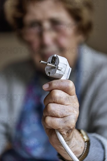 Senior citizen holding a power cable with plug in her hand at home, symbolising energy costs and poverty, Cologne, North Rhine-Westphalia, Germany, Europe