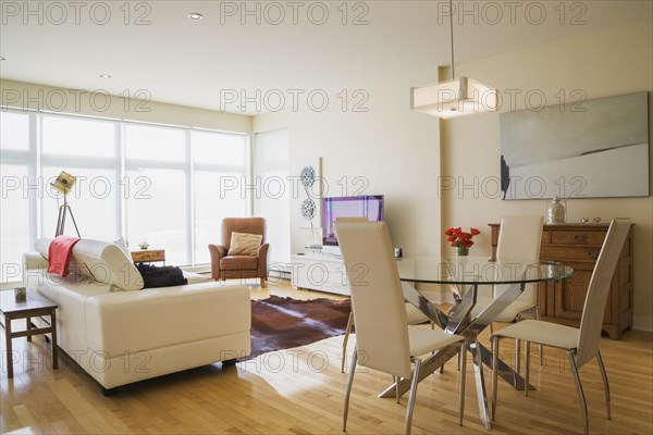Dining room area with round glass table, white high-back leather chairs and white leather sofa in living room inside a renovated ground floor apartment in an old residential cottage style home, Quebec, Canada, North America