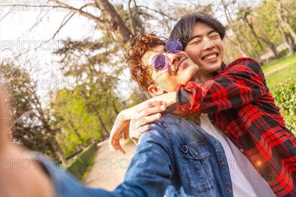 Multicultural smiling and cute gay couple taking selfie embracing in a park