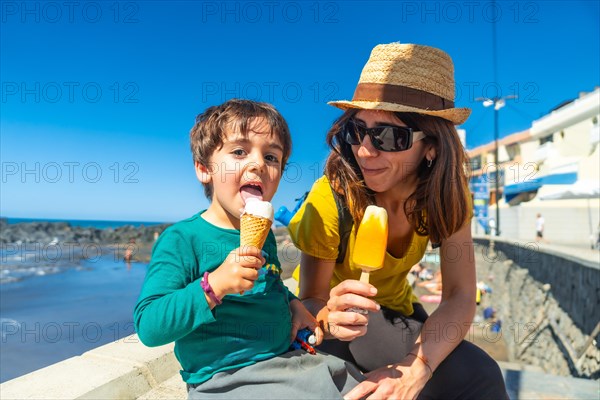 A woman and a child are sitting on a wall near the ocean, eating ice cream. The woman is wearing a straw hat and sunglasses