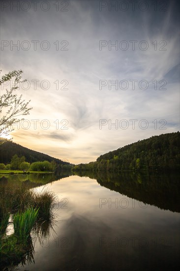 A lake in a landscape shot. A sunset and the natural surroundings are reflected in the water of the reservoir. Marbach reservoir, Odenwald, Hesse