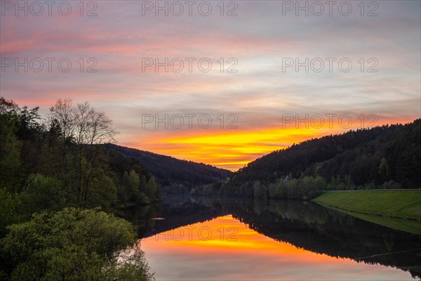 A lake in a landscape shot. A sunset and the natural surroundings are reflected in the water of the reservoir. Marbach reservoir, Odenwald, Hesse