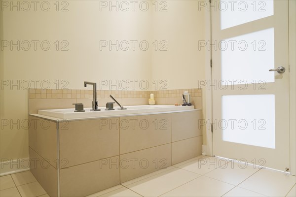 Soaking tub encased in a ceramic tile base in bathroom inside a renovated ground floor apartment in an old residential cottage style home, Quebec, Canada, North America