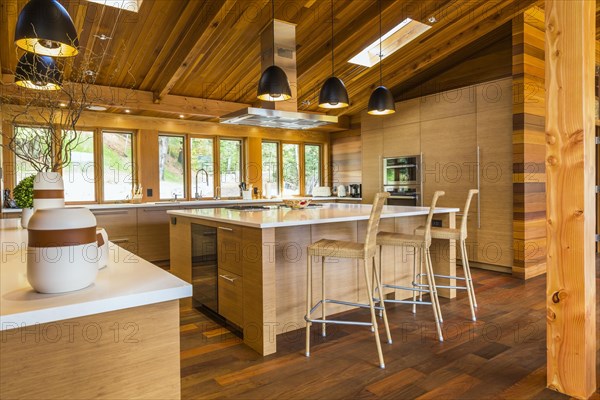 Bamboo wood buffet and island with white quartz countertops and high wicker chairs, cabinets in kitchen with Ipe wood floor plus black industrial style pendant lighting fixtures inside luxurious stained cedar and timber wood home with panoramic windows, Quebec, Canada, North America