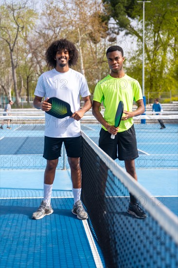 Vertical Portrait of two young african american pickleball players standing looking at camera holding pickleball equipment