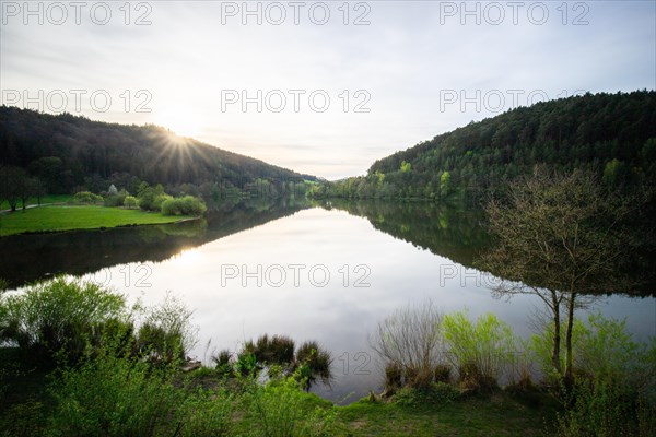 A lake in a landscape shot. A sunset and the natural surroundings are reflected in the water of the reservoir. Marbach reservoir, Odenwald, Hesse