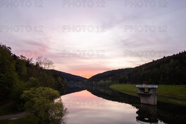 A lake in a landscape shot. A sunset and the natural surroundings are reflected in the water of the reservoir. Marbach reservoir, Odenwald, Hesse