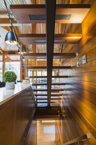 Underside view of illuminated wooden staircase with opened steps and clear glass railing inside luxurious stained cedar and timber wood home, Quebec, Canada, North America